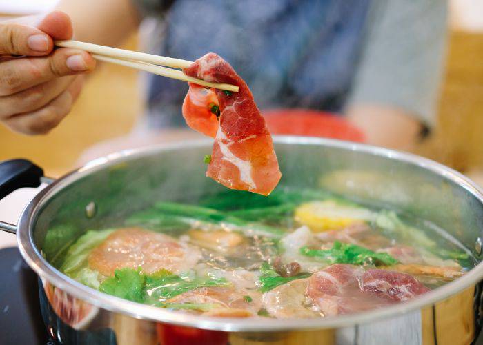 Someone preparing to dunk a thin slice of meat into a bubbling hot pot broth.